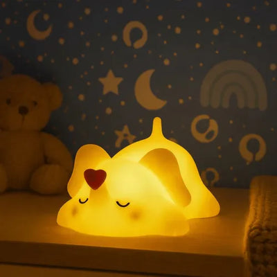 Veilleuse éléphant bébé en silicone avec cœur rouge allumée diffusant une lumière chaude ambrée sur une étagère de chambre d'enfant, à côté d'une peluche ourson, avec un papier peint étoiles et lunes en arrière-plan créant une ambiance nocturne apaisante