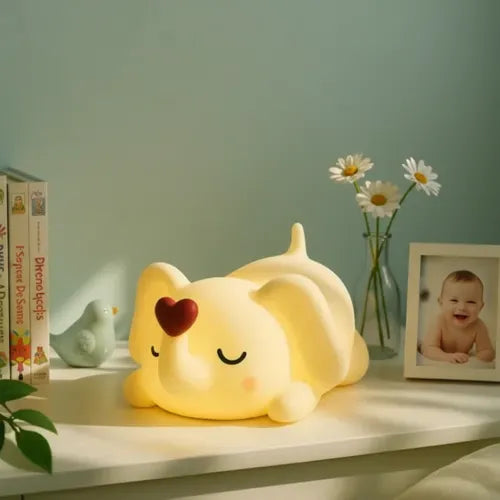 Veilleuse éléphant bébé en silicone souple jaune avec petit cœur rouge, lumière LED tamisée et chaleureuse, posée sur une étagère de chambre d'enfant décorée de livres, fleurs et cadres photo